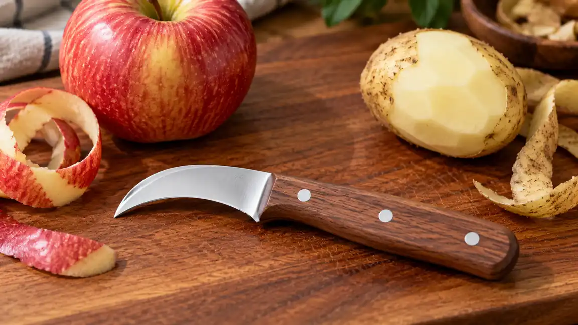 Bird's-beak paring knife with a curved blade beside an apple and a turned potato.